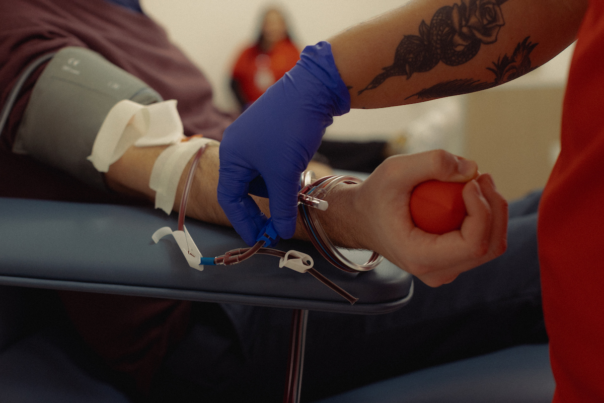 Blood donor squeezes red ball while a phlebotomist tapes the tubes down