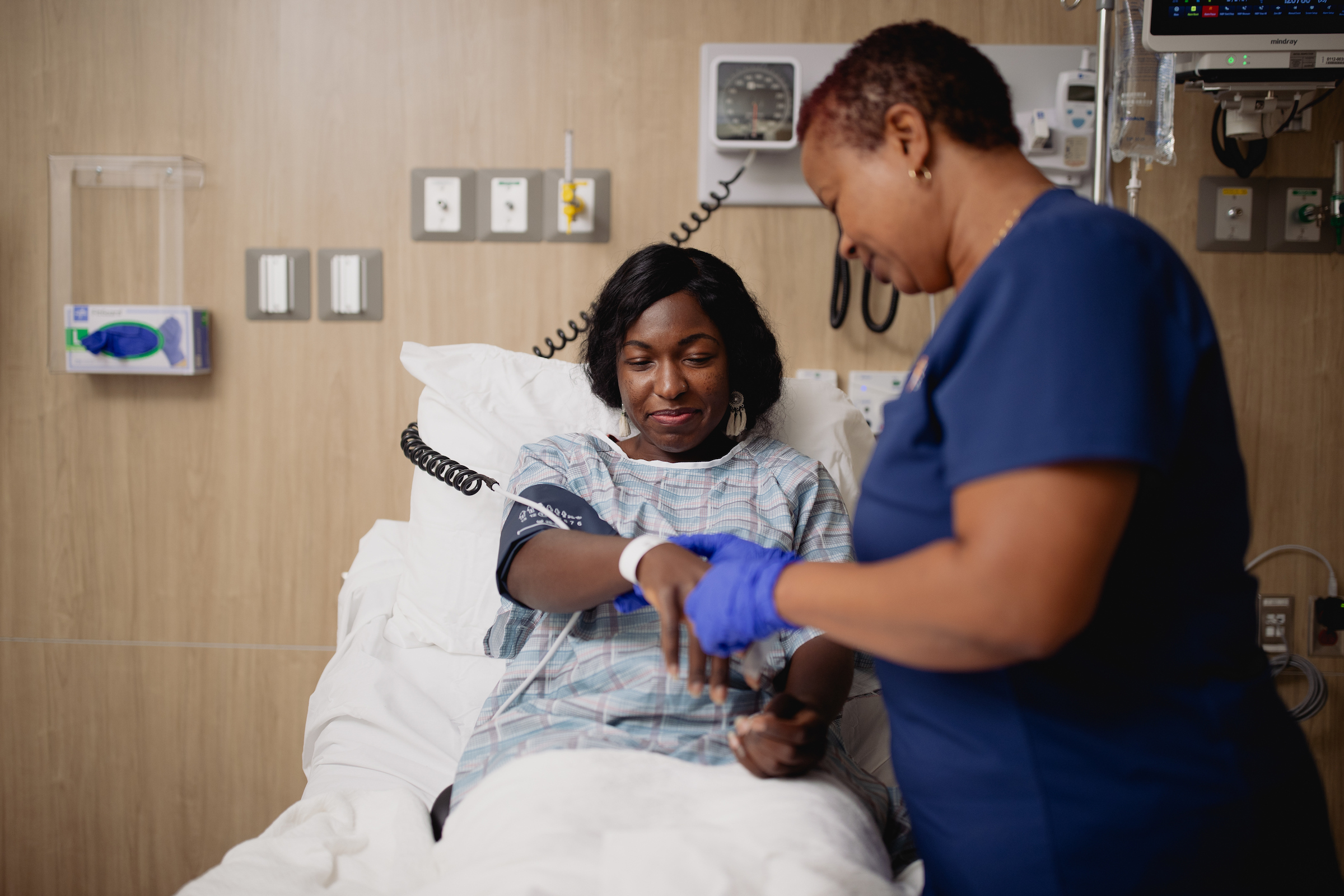 Patient in a hospital bed smiles while a nurse assists with monitoring equipment, highlighting supportive, hands-on care during recovery.