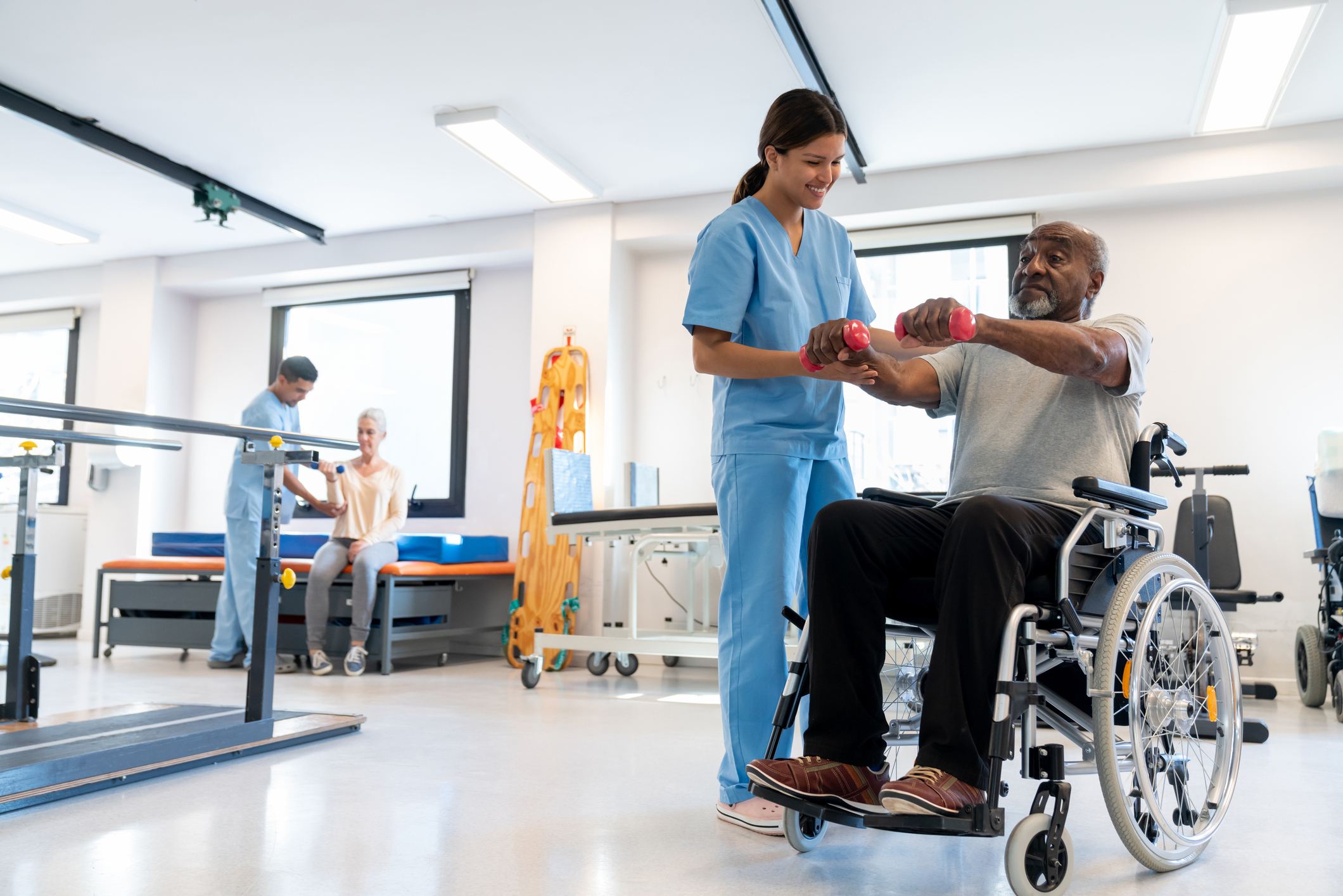 Cheerful therapist correcting the posture of her black senior patient on a wheelchair working out his arms with dumbbells